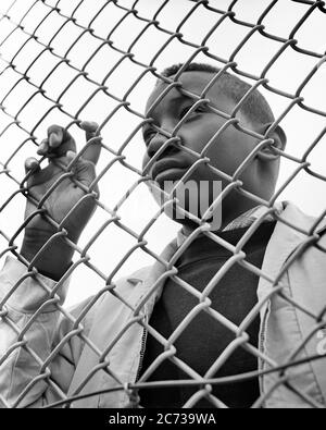 1960s AFRICAN-AMERICAN TEENAGE BOY LOOKING THRU A CHAIN-LINK FENCE - n1932 HAR001 HARS FREEDOM GOALS DREAMS AFRICAN-AMERICANS AFRICAN-AMERICAN LOW ANGLE BLACK ETHNICITY CONCEPTUAL DEPRIVED ESCAPE TEENAGED DENIED DISADVANTAGED LOOKING IN BARRIER CHAIN-LINK CONFINED DISAFFECTED DISAPPOINTED DISCONNECTED GROWTH IMPOVERISHED JUVENILES TRAPPED BLACK AND WHITE DISTRAUGHT HAR001 OLD FASHIONED RESTRICT THRU AFRICAN AMERICANS Stock Photo