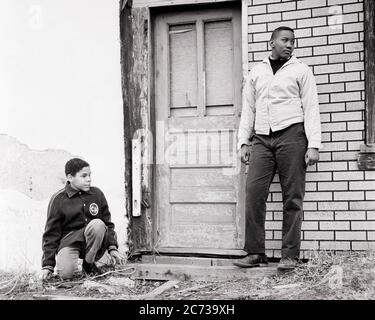 1960s TWO BORED AFRICAN AMERICAN TEENAGE BOYS STANDING BY DOOR TO ABANDONED BUILDING IN POVERTY STRICKEN URBAN NEIGHBORHOOD - n1956 HAR001 HARS BROTHERS POOR HOME LIFE COPY SPACE FRIENDSHIP FULL-LENGTH PERSONS DANGER MALES RISK TEENAGE BOY SIBLINGS B&W SADNESS FREEDOM TEMPTATION DREAMS NEIGHBORHOOD STRENGTH AFRICAN-AMERICANS AFRICAN-AMERICAN CHOICE POWERFUL BLACK ETHNICITY DIRECTION PRIDE BY IN TO ABANDONED SIBLING CONNECTION CONCEPTUAL ESCAPE SUPPORT TEENAGED DISADVANTAGED CAUTION DISAFFECTED DISAPPOINTED DISCONNECTED IDEAS IMPOVERISHED JUVENILES PRE-TEEN BOY STRICKEN TOGETHERNESS Stock Photo