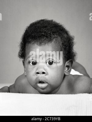 1960s AFRICAN-AMERICAN BABY LAYING ON STOMACH FUNNY FACIAL EXPRESSION - n2307 HAR001 HARS AFRICAN-AMERICANS AFRICAN-AMERICAN BLACK ETHNICITY COMICAL COMEDY BABY BOY WIDE-EYED GROWTH JUVENILES BIG EYES BLACK AND WHITE HAR001 MOUTH OPEN OLD FASHIONED AFRICAN AMERICANS Stock Photo