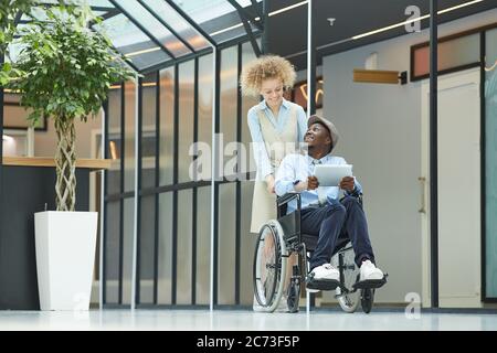 Young woman walking along the corridor with disabled man in wheelchair he using tablet pc Stock Photo