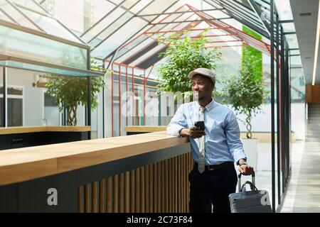 African young businessman with suitcase using his mobile phone while standing in the waiting room Stock Photo