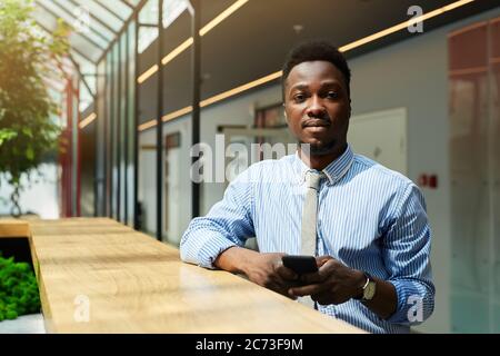 Portrait of African young businessman standing with mobile phone and looking at camera at office Stock Photo