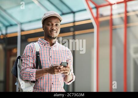 African young man in cap with backpack behind his back smiling at camera while using his mobile phone Stock Photo