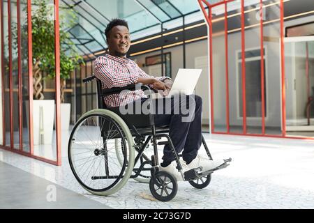 Portrait of African disabled man sitting in wheelchair and looking at camera while working on laptop Stock Photo