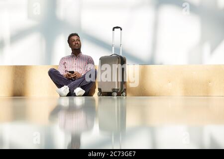 African man sitting on the floor with suitcase and using his mobile phone at the airport Stock Photo
