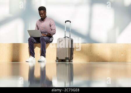African young businessman sitting at the airport with luggage and working on laptop while waiting for his flight Stock Photo