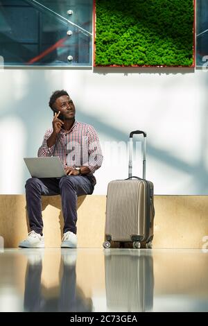 African young businessman talking on mobile phone and uisng laptop while sitting at the airport he ordering plane tickets Stock Photo