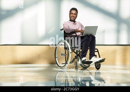 Portrait of African disabled man in wheelchair looking at camera while working online on laptop computer Stock Photo