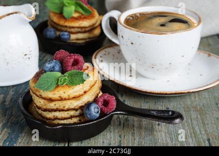 Breakfast of pancakes in a portioned cast-iron frying pan, fresh berries and black coffee in a vintage ceramic cup on an old wooden table Stock Photo