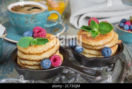 Breakfast of pancakes in cast-iron frying pans, fresh berries and black coffee on old wooden table, in rustic style Stock Photo