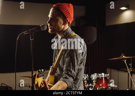 young caucasian gorgeous musician play electric guitar and sing on microphone in recording studio. rockstar perform music. man preparing, practicing b Stock Photo