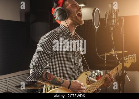 young caucasian gorgeous musician play electric guitar and sing on microphone in recording studio. rockstar perform music. man preparing, practicing b Stock Photo