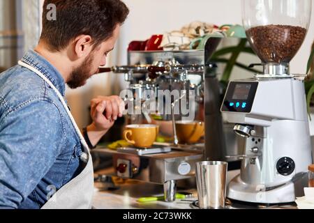Caucasian young barman with beard, stands near coffee machine with back to camera, holds tamper, press cofee, prepares americano at bar counter Stock Photo