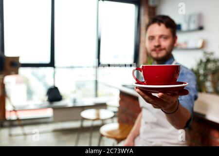 Bearded hipster barman, wears white apron, stretches hand towards camera, keeps big beautiful coffee set, blurred background, small business concept Stock Photo