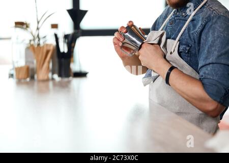 Hands of solid man wearing white neat apron holding towel, cleaning steel pitcher in brightly lighted room, background of window in modern restaurant Stock Photo