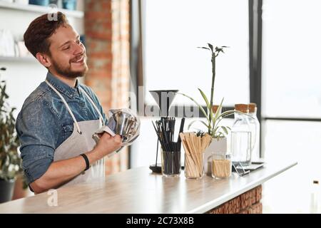 Delighted young male barista in neat apron, looks kindly away with broad smile, keeps towel and Turkish coffee pot, background of window Stock Photo