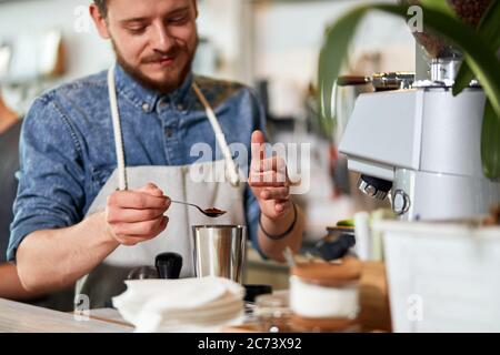 Handsome peaceful male barista stands in apron behind counter, keeps small spoon with instant coffee, expresses satisfaction, looks away with smiling Stock Photo