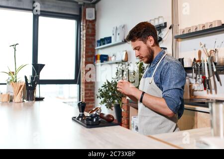 Young handsome barista dre sed in casual outfit and neat apron, works at light coloured counter, focuses on cover in hands, tamper with coffee in fron Stock Photo