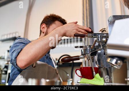 Young bearded man stands near professional grinder, keeps handle, watches coffee pouring process, prepares fresh aromatic cappuccino for customer in m Stock Photo