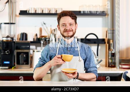 Positive peaceful barman dressed in jeans shirt and wgite apron, stands kindly at counter, looks straight at camera with glad face, smiles friendle, k Stock Photo