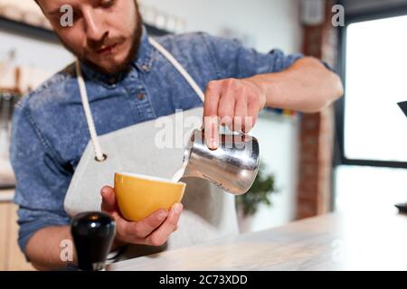 Confident concentrated bartender in neat apron pours steamed hot milk from metal jug into brightly coloured small coffee cup, work process concept Stock Photo