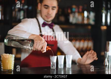 Expert barman in elegant uniform working at night party at popular nightclub, pouring vodka in frosted short glasses Stock Photo