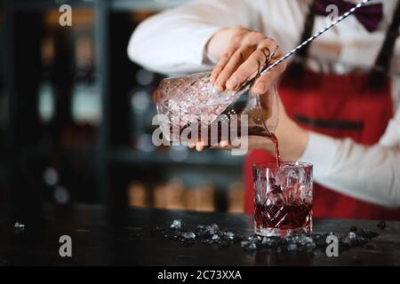 The bartender man pours ruby cocktail into crystal wide glass, close up. Bar, Restaurant, Bartendering, Beverage and Service concept. Stock Photo