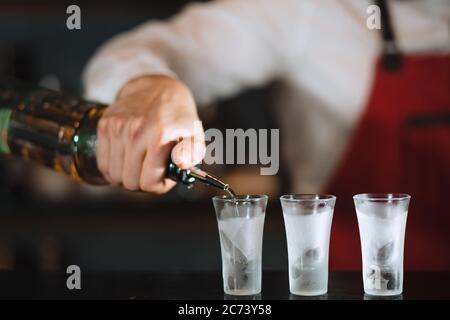 Close up photo of hands of a bartender pouring some drink from the bottle into shot frozen glasses on a wooden counter. Stock Photo