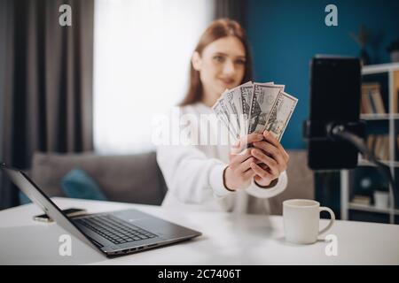 Successful blogger with brown hair showing on camera dollars banknotes while sitting at table. Attractive girl explaining her followers in social netw Stock Photo