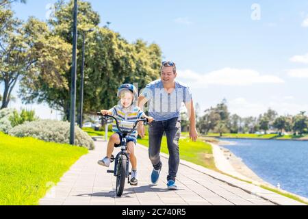 Boy learning to ride a bicycle with his father in the park by the lake. Father and son having fun together on the bikes. Happy family, outdoors activi Stock Photo