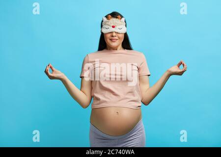 Pregnant woman wearing sleep mask on eyes meditates, keeping fingers in mudra while standing at yoga studio. Woman enjoying in meditation. Stock Photo