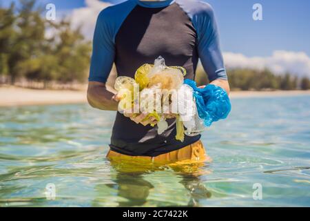 Man collects packages from the beautiful turquoise sea. Paradise beach pollution. Problem of spilled rubbish trash garbage on the beach sand caused by Stock Photo