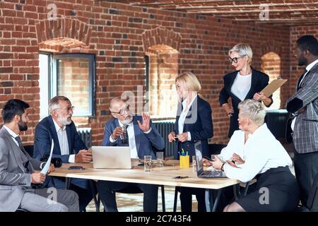 Four businessmen wear grey and blue suits, and four blonde woman wear office clothes discuss trade at table in office Stock Photo