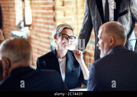 Focused on blonde caucasian woman in glasses smile, talk between backs of two men Stock Photo