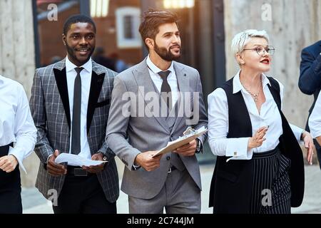 Three businessmen move outdoor. African man in checked jacket and caucasian man in grey jacket keep papier. Blonde caucasian woman move energetically. Stock Photo