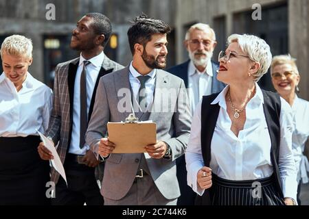 Group of busy business people in differnt suits walking down the street. In the foreground brunette bearded man looks at blonde in white shirt. in the Stock Photo