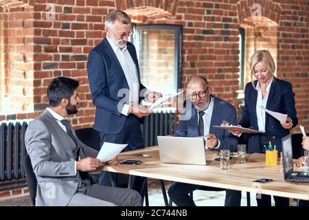 Four adult caucasian wear stylish office clothes look at notes at table in meeting room. On table phones. laptops, glasses of water Stock Photo