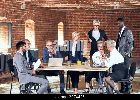 Four businessmen wear grey and blue suits, and four blonde woman wear office clothes actively discuss trade at table in office Stock Photo