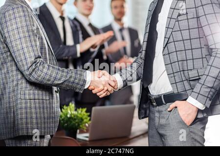close-up of business people, young guys shaking hands, wearing grey elegant tuxedo, happy men colleagues applause to them in the background Stock Photo