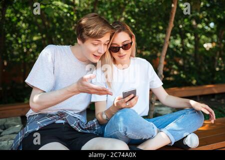 Joyful couple sitting on bench in park and emotionally discussion something while using cellphone. Close up photo of young smiling man and nice lady Stock Photo