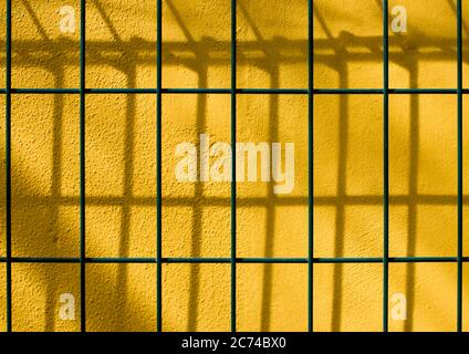 Close up of yellow wall behind the green fence mesh. Yellow color wall background. Texture of wire mesh fence. Stock Photo