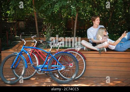 Close up of red and blue bicycles in park. Boy sitting on bench in park with pretty girl with blond hair that leaning on him and taking selfie Stock Photo