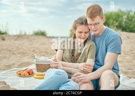 Young couple enjoying picnic on beach embracing and holding cups Stock Photo