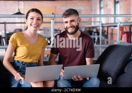 positive businessman and businesswoman holding laptops while sitting on sofa Stock Photo