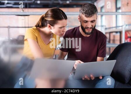 selective focus of businessman and emotional businesswoman using laptops while sitting on sofa Stock Photo