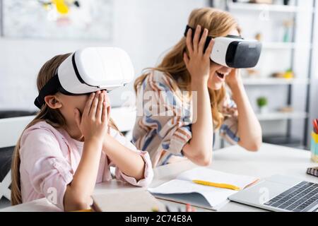 Selective focus of excited kid covering mouth while using vr headsets with mother near laptop and stationery Stock Photo