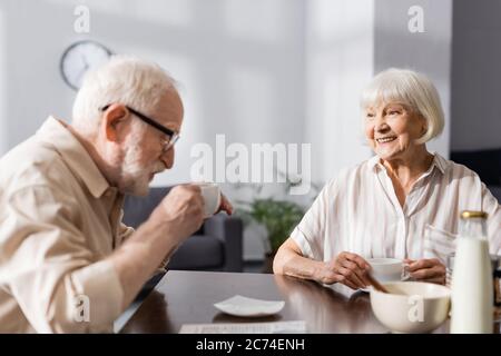 Selective focus of smiling senior woman looking at husband drinking coffee at home Stock Photo