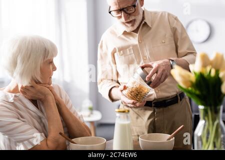 Selective focus of senior man opening jar with cereals near wife during breakfast in kitchen Stock Photo
