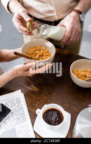 Cropped view of woman holding bowl with cereals while husband pouring milk near coffee, newspaper and smartphone on table Stock Photo