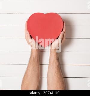 Male hands hold a red heart shaped box against the background of a white wooden table. Stock Photo